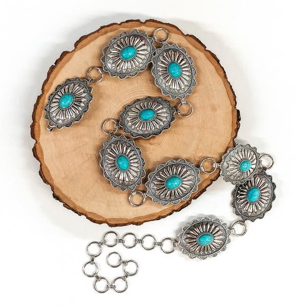 Silver concho belt with turquoise center stones. This belt is pictured on a white background laid on a piece of wood. 