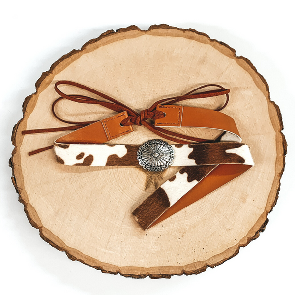 Brown cow print hat band with a single silver stone cluster. It also has leather like material strands that tie it together. It is laying on a piece of wood that is pictured of a white background.