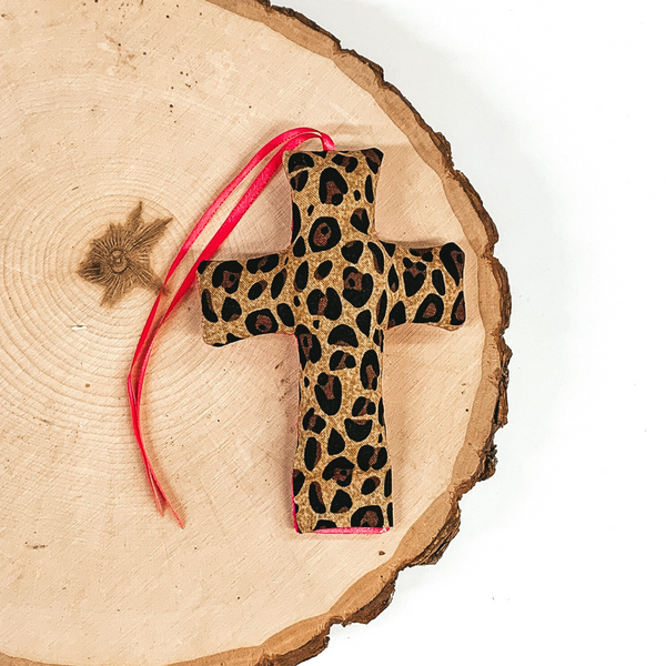 Cross shaped freshie that is leopard print with pink string. This is laying on a piece of wood that is on a white background.