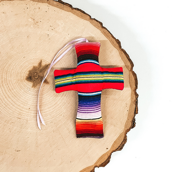 Cross shaped freshie that is serape print with red string. This is laying on a piece of wood that is on a white background.