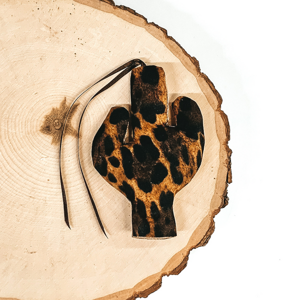 Cactus shaped freshie that is a leopard print with brown string. This is laying on a piece of wood that is on a white background.