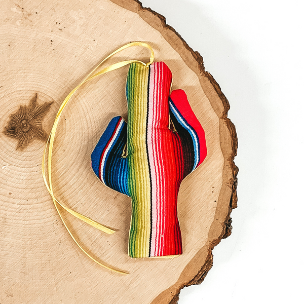 Cactus shaped freshie that is a serape print with yellow string. This is laying on a piece of wood that is on a white background.