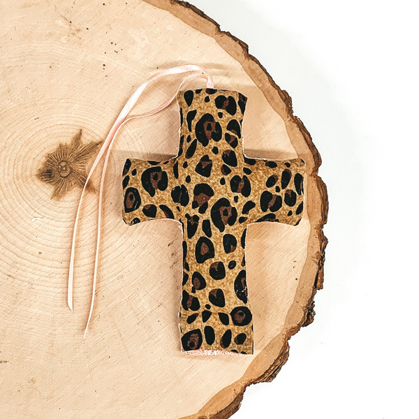 Cross shaped freshie that is a leopard print with light pink string. This is laying on a piece of wood that is on a white background.