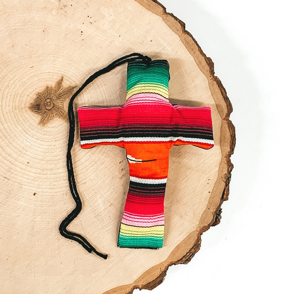 Cross shaped freshie that is a serape print with black string. This is laying on a piece of wood that is on a white background.