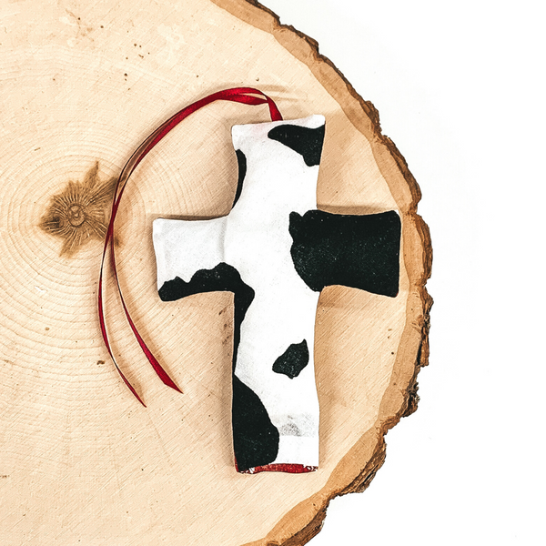 Cross shaped freshie that is a black and white cow print with red string. This is laying on a piece of wood that is on a white background.