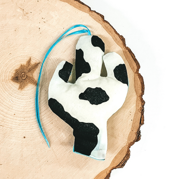 Cactus shaped freshie that is a black and white cow print with light blue string. This is laying on a piece of wood that is on a white background.
