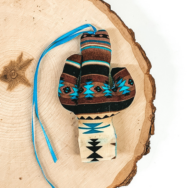 Cactus shaped freshie that is a brown and turquoise aztec print with blue string. This is laying on a piece of wood that is on a white background.