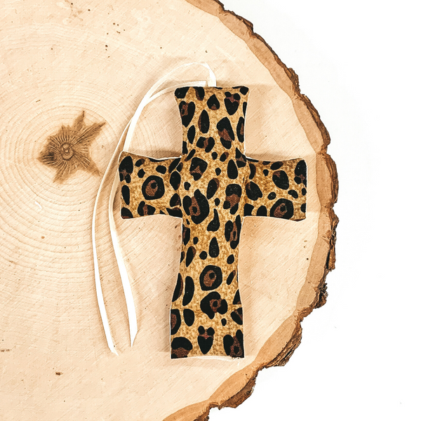 Cross shaped freshie that is a leopard print with white string. This is laying on a piece of wood that is on a white background.