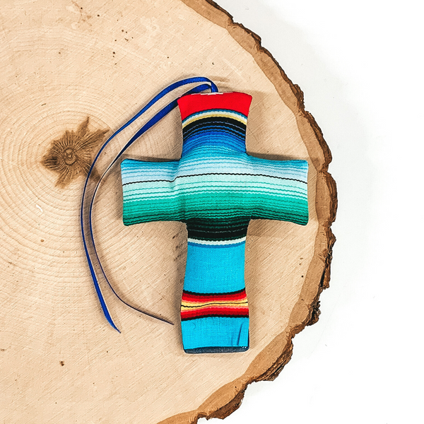 Cross shaped freshie that is a serape print with blue string. This is laying on a piece of wood that is on a white background.