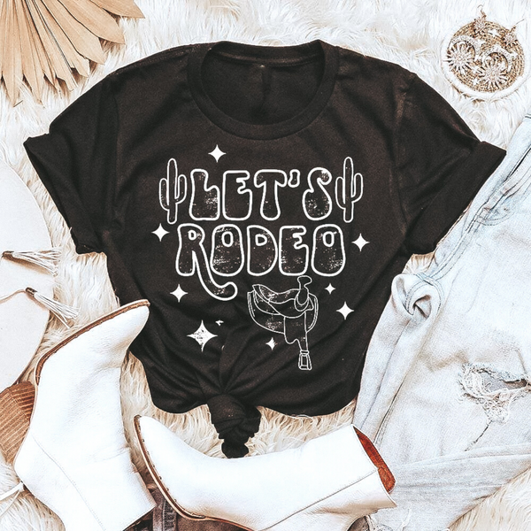 Black graphic tee with "Let's Rodeo" in cute font.  Surrounded by stars, cacti and saddle.  You can see this tee paired with white booties and light wash jeans.  Shirt is laid on top of a fuzzy run with cute trinkets surrounding.
