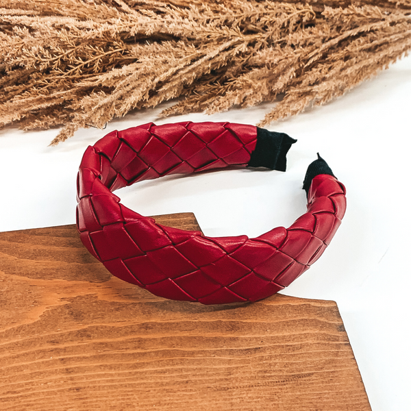 This headband has a red basket weave or braided pattern around the entire headband. This headband is pictured laying in a piece of wood with tan floral at the top of the picture on a white background.