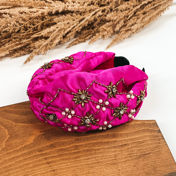 This is a thick, fuchsia colored headband with gold and pearl beaded design. This headband is pictured laying in a piece of wood with tan floral at the top of the picture on a white background.