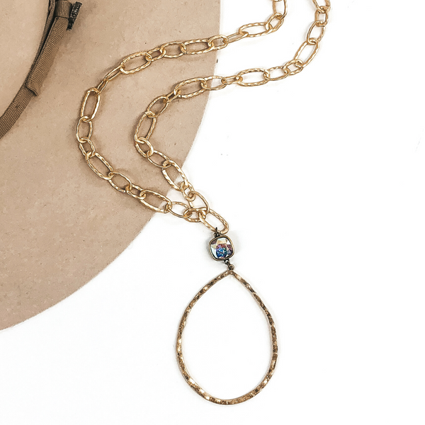 Gold chain link necklace with a open hammered teardrop pendant with a ab cushion cut crystal connector. This necklace is laying partially on a beige hat that is on a white background. 