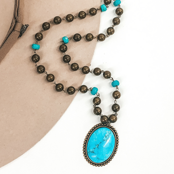 Bronze and turquoise beaded necklace with a bronze oval pendant with a turquoise cabochon stone. This is pictured on a white and beige background.