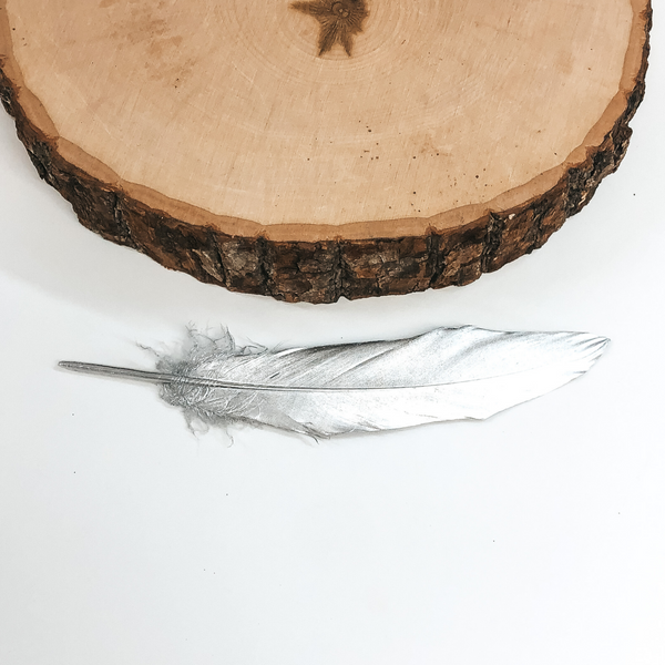 This is a metallic silver colored feather. This feather is pictured on a white background with a piece of wood at the top of the picture. 