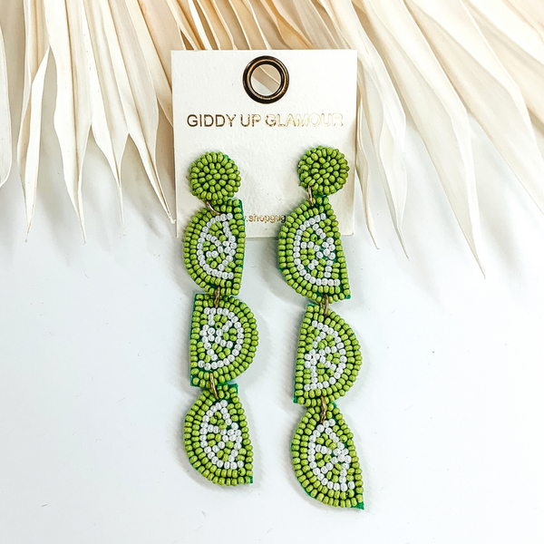 These are lime green beaded earrings. It is in the shape of three halved limes connected together. These earrings are pictured laying partially on some ivory leaves and on a white background. 