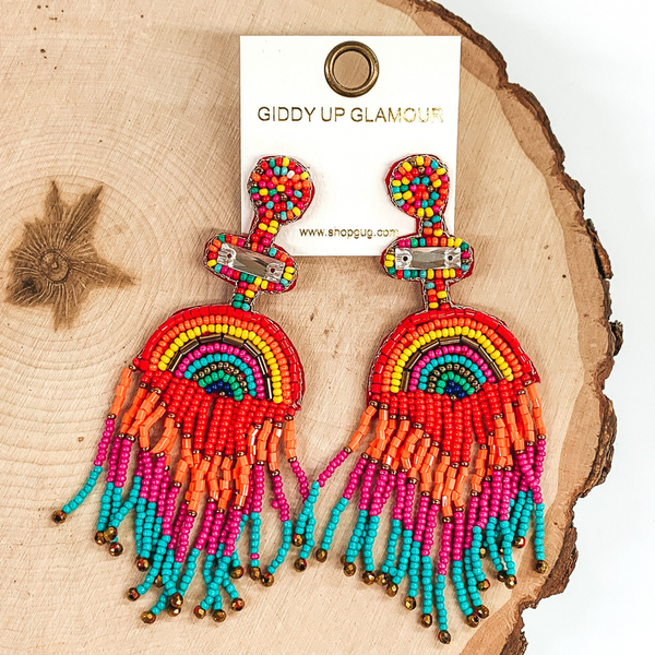 These are a multicolored beaded earrings with multicolored tassels. There is also a clear, rectangle, crystal bead towards the top of the earrings. these earrings are pictured laying on a piece of wood on a white background. 