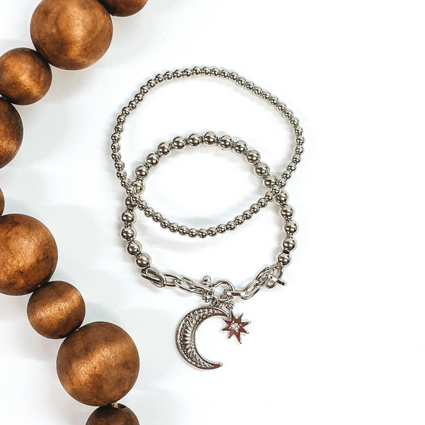 This silver bracelet set includes two beaded bracelets. One is a plain beaded bracelet and the other one has a chained segment with a moon and star charm. This bracelet set is pictured on a white background with brown beads on the side of the picture.