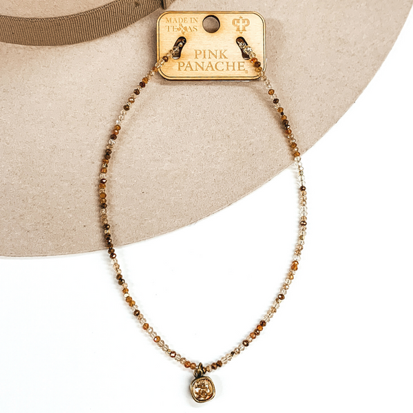 Rose gold and bronze mix beaded adjustable necklace with a single square cushion cut crystal in the color topaz. This necklace is pictured laying on a white and beige background. 