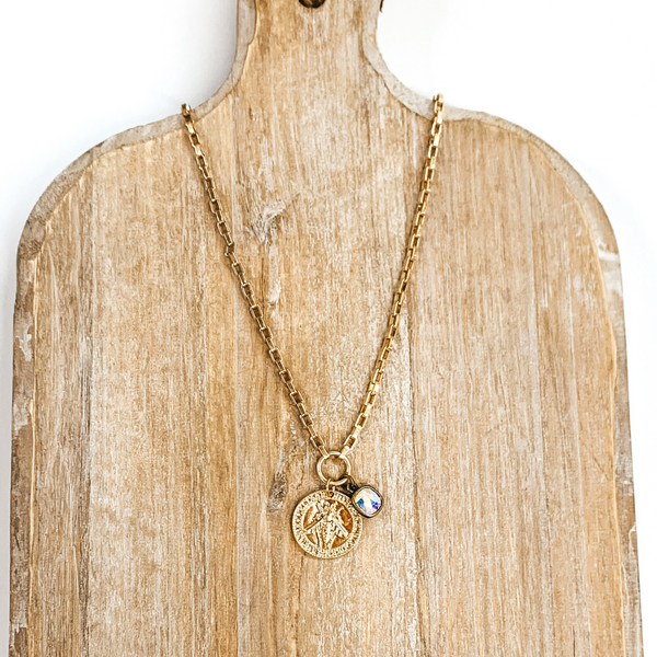 Gold chained necklace with a hanging gold coin pendant and hanging ab crystal. On the coin there is an engraved bee. This necklace is pictured on a tan necklace holder and on a white background. 