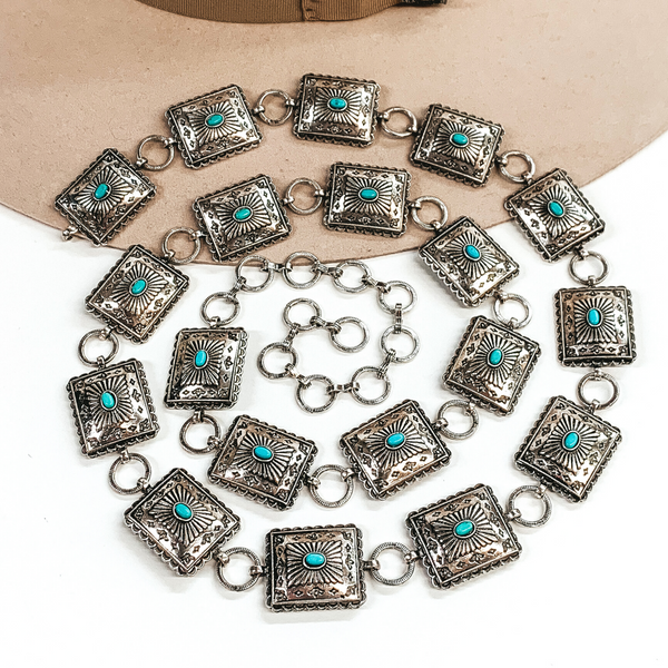 Silver square concho belt with tiny center turquoise stones. This belt also includes silver circle loops to make it adjustable. This belt is pictured laying partially on a beige colored hat on a white background. 