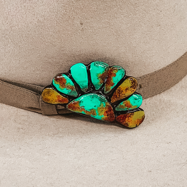 This hat pin is a half of a concho. The main color is turquoise and is mixed with brown, red and yellow like real turquoise. This hat pin is pictured in front of a beige hat band. 