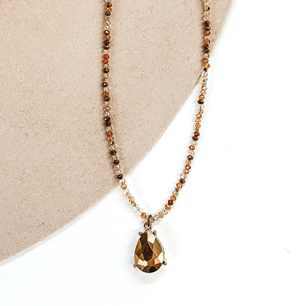 Bronze and rose gold mix of crystal beads necklace with a hanging teardrop pendant. The pendant is a gold metallic color. This necklace is pictured on a white and beige background. 