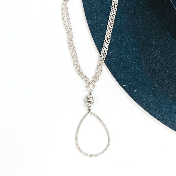 Double layered silver chained necklace with front, small toggle clasp. This necklace includes a hanging clear cushion cut crystal and an open teardrop silver pendant. This necklace is pictured on a navy and white background. 