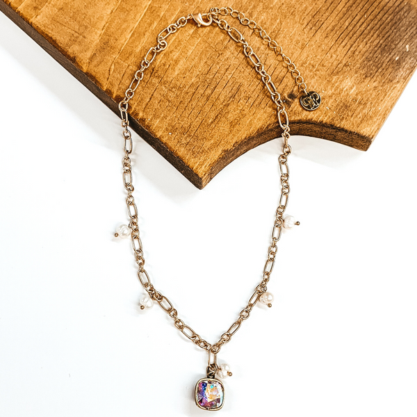 Gold chained necklace with five dangle, white pearls and a hanging ab cushion cut crystal. This necklace is partially laying on a dark piece of wood on a white background.