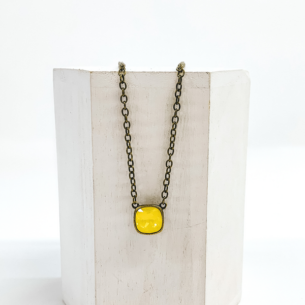 Bronze chained necklace with a square pendant with a yellow cushion cut crystal. This necklace is pictured laying on a white block on a white background. 