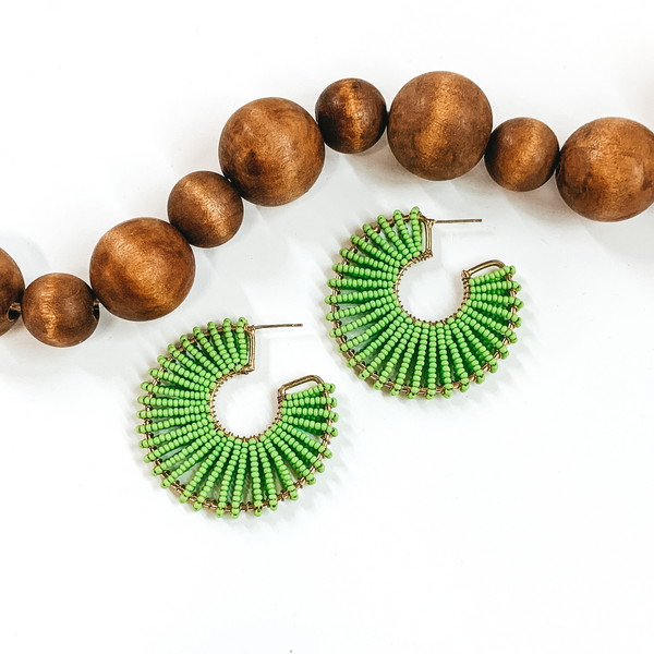 Gold outlined, thick hoops. Wrapped around the gold outline are a lime green beaded string. These earrings are pictured on a white background with dark brown beads at the top of the picture. 