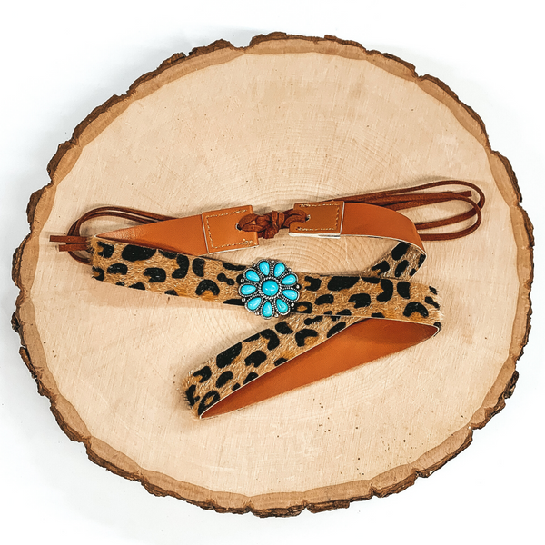 Leopard hat band with a single turquoise stone cluster concho. It also has leather like material strands that tie it together. It is laying on a piece of wood that is pictured of a white background.