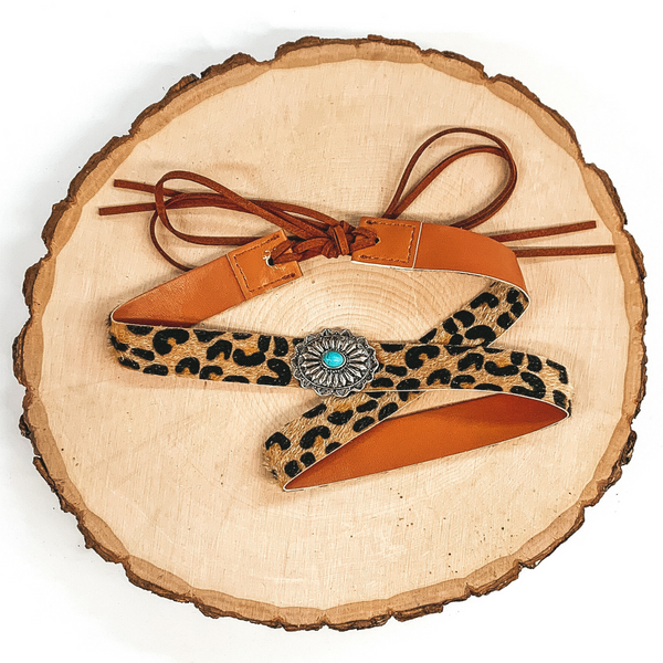 Leopard hat band with a single silver concho that has a turquoise center stone. It also has leather like material strands that tie it together. It is laying on a piece of wood that is pictured of a white background.
