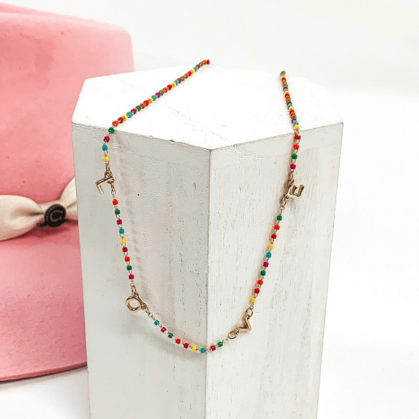 This is a multicolored beaded necklace with gold letter charms that spell out "LOVE." This necklace is pictured on a white block on a white background with a pink hat in the background. 