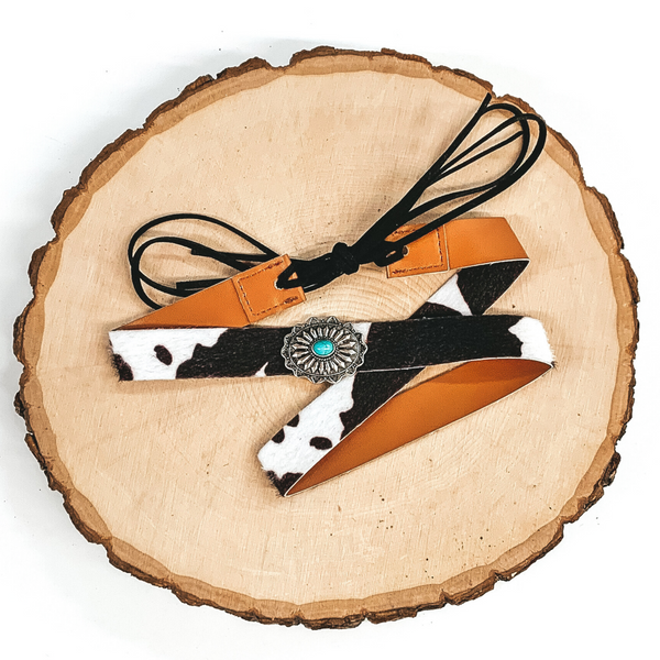 Black cow print hat band with a single silver concho with a turquoise center stone. It also has leather like material strands that tie it together. It is laying on a piece of wood that is pictured of a white background.