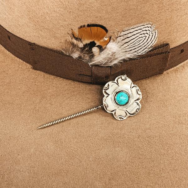 Silver twisted toothpick hat pin with a silver flower pendant. This flower has small engravings and a center turquoise colored stone. This hat pin is pictured on a tan hat.