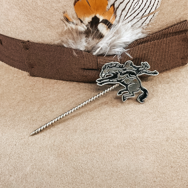 Silver twisted toothpick hat pin with a silver pendant a cowboy on a horse. The horse is bucking while the cowboy is on top with one hand in the air and one on the reins. This hat pin is pictured on a tan hat.