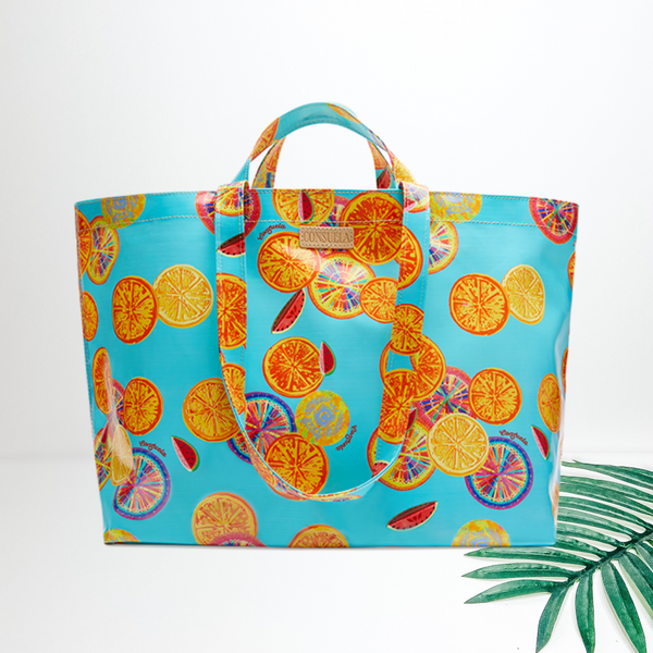 A large size blue bag with a colorful fruit pattern. Pictured on white background with a straw hat and palm leaf.