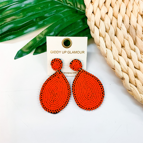 Circle, beaded post back stud earrings with a hanging, teardrop beaded pendant in red. These earrings are pictured on a tan basket weave with a green leaf in the background. 