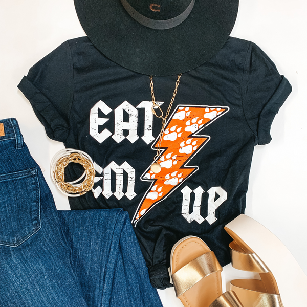 A black graphic tee that says "Eat Em Up Kats" with an orange lightning bolt. Pictured on a white background with black jeans, gold wedges, and gold jewelry.