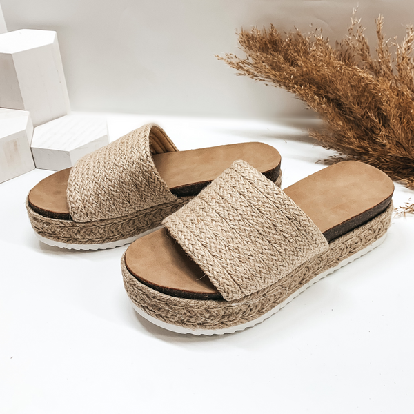A pair of platform espadrille sandals with a slide on upper. Pictured on white background with pampas grass.