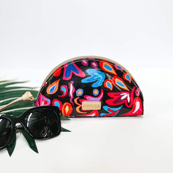 A black dome shaped makeup bag that has a colorful swirly print. Pictured on white background with black sunglasses and a palm leaf.