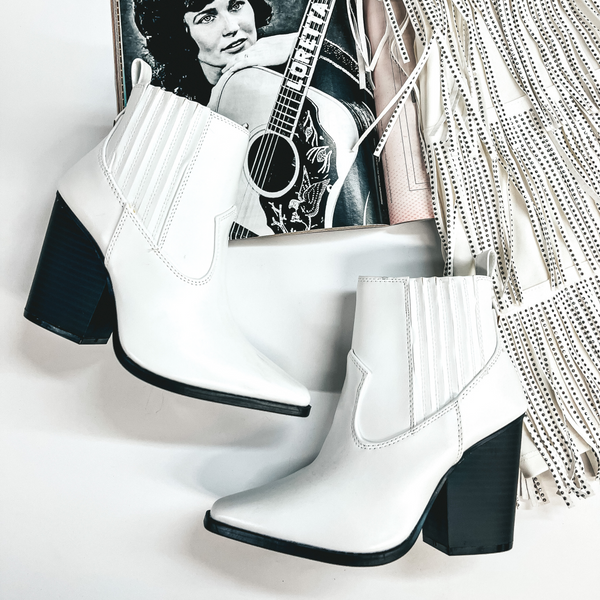 A pair of white booties with a black sole and high heel. These western booties are pictured on a white background with crystal fringe and a magazine.