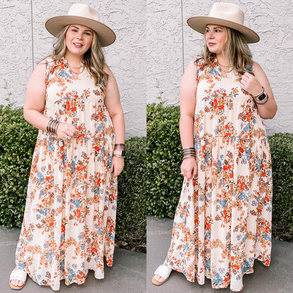 Model is wearing an ivory maxi dress with an orange and blue floral print. Model has it paired with a beige hat, silver jewelry, and ivory sandals.