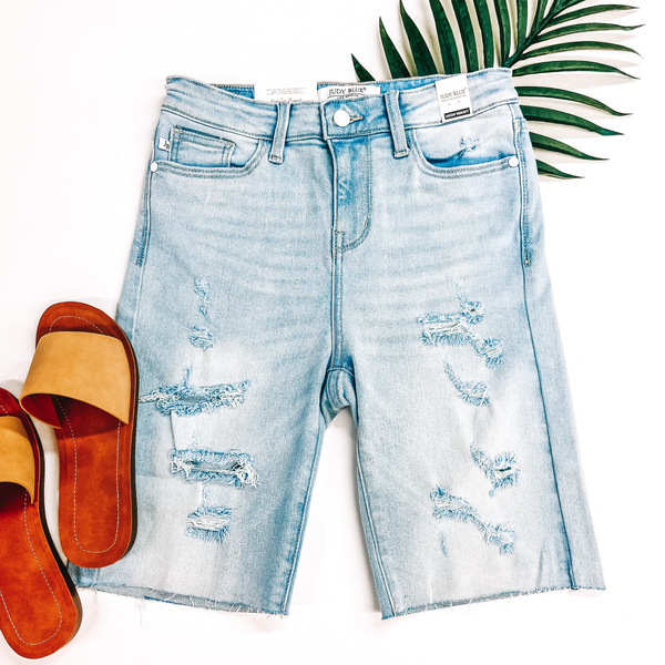 A pair of distressed Bermuda shorts in light blue. These shorts are pictured on a white background with yellow sandals and a palm leaf.