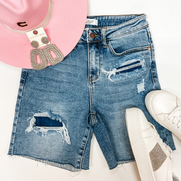 A pair of patch distressed mid thigh shorts. Pictured on white background with crystal earrings, a pink hat, and crystal sneakers.