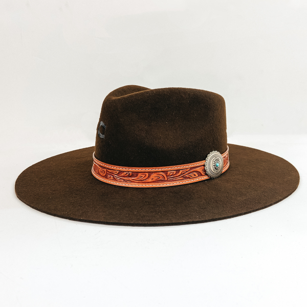 A chocolate brown felt hat with a leather tooled band with a silver concho pin. Pictured on white background.