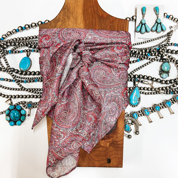 A pink and purple paisley print wild rag tied around a wooden display. Pictured on white background with silver and turquoise jewelry.