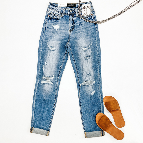 A pair of medium wash boyfriend jeans with distressing on the legs and cuffed ankles. Pictured on a white background with tan sandals and sterling silver jewelry.