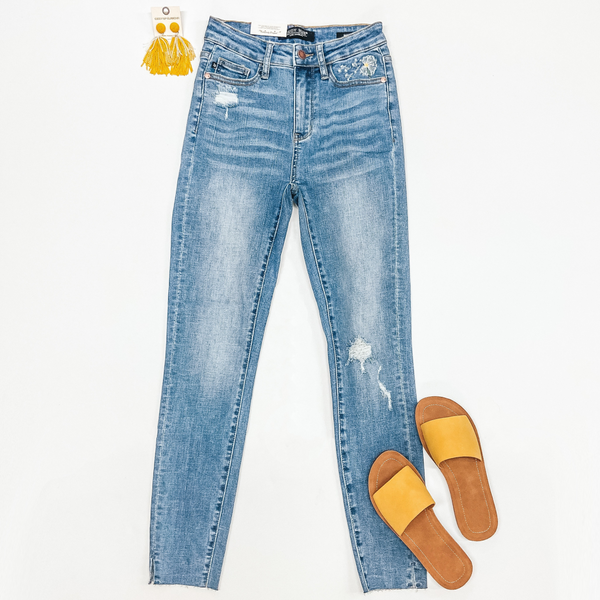 A pair of medium wash skinny jeans with dandelion embroidery and distressing. These jeans are pictured on a white background with yellow sandals and yellow earirngs.